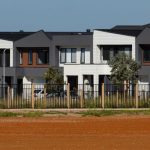 Modern townhouses with neutral-toned exteriors, a white fence and mature trees lining a quiet residential street.
