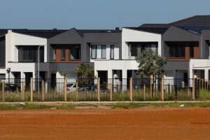 Modern townhouses with neutral-toned exteriors, a white fence and mature trees lining a quiet residential street.