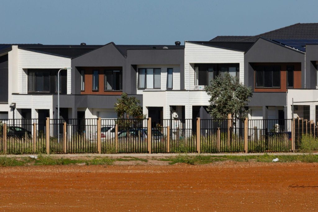 Modern townhouses with neutral-toned exteriors, a white fence and mature trees lining a quiet residential street.