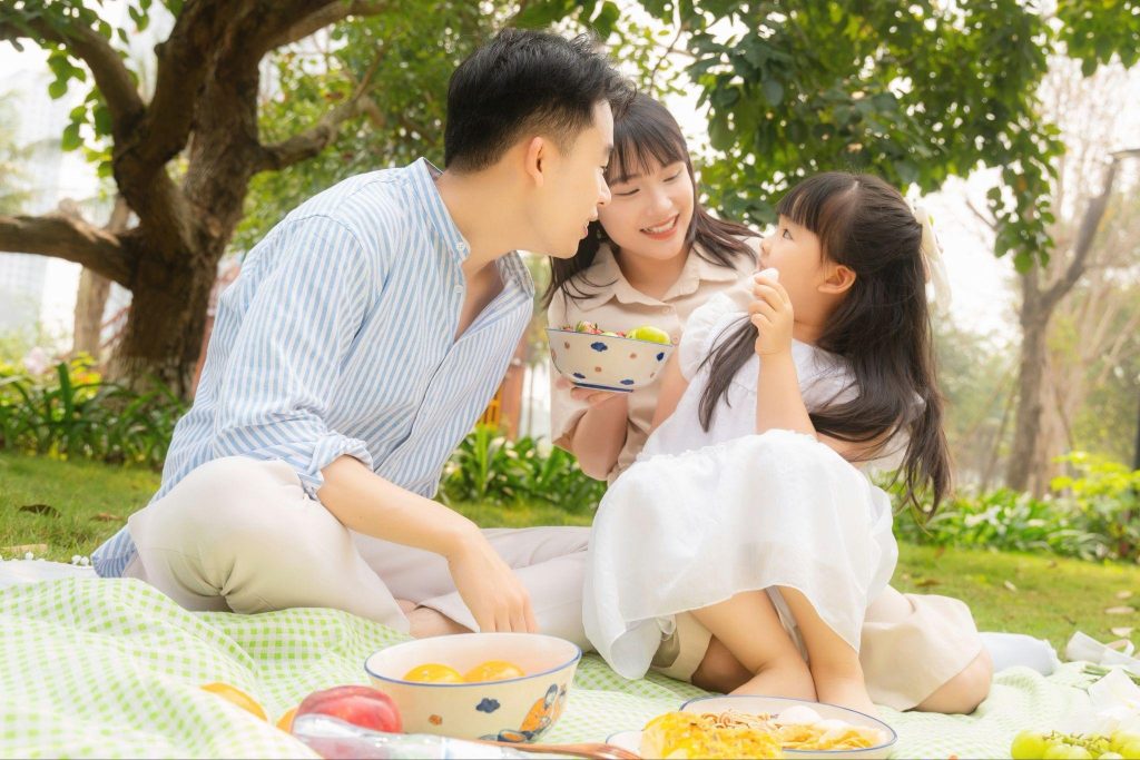 A family enjoying a picnic together on a blanket in a sunny park, with food and fruit spread out in front of them.