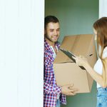 Young couple packing boxes at a home doorway, woman holding a clipboard checklist.