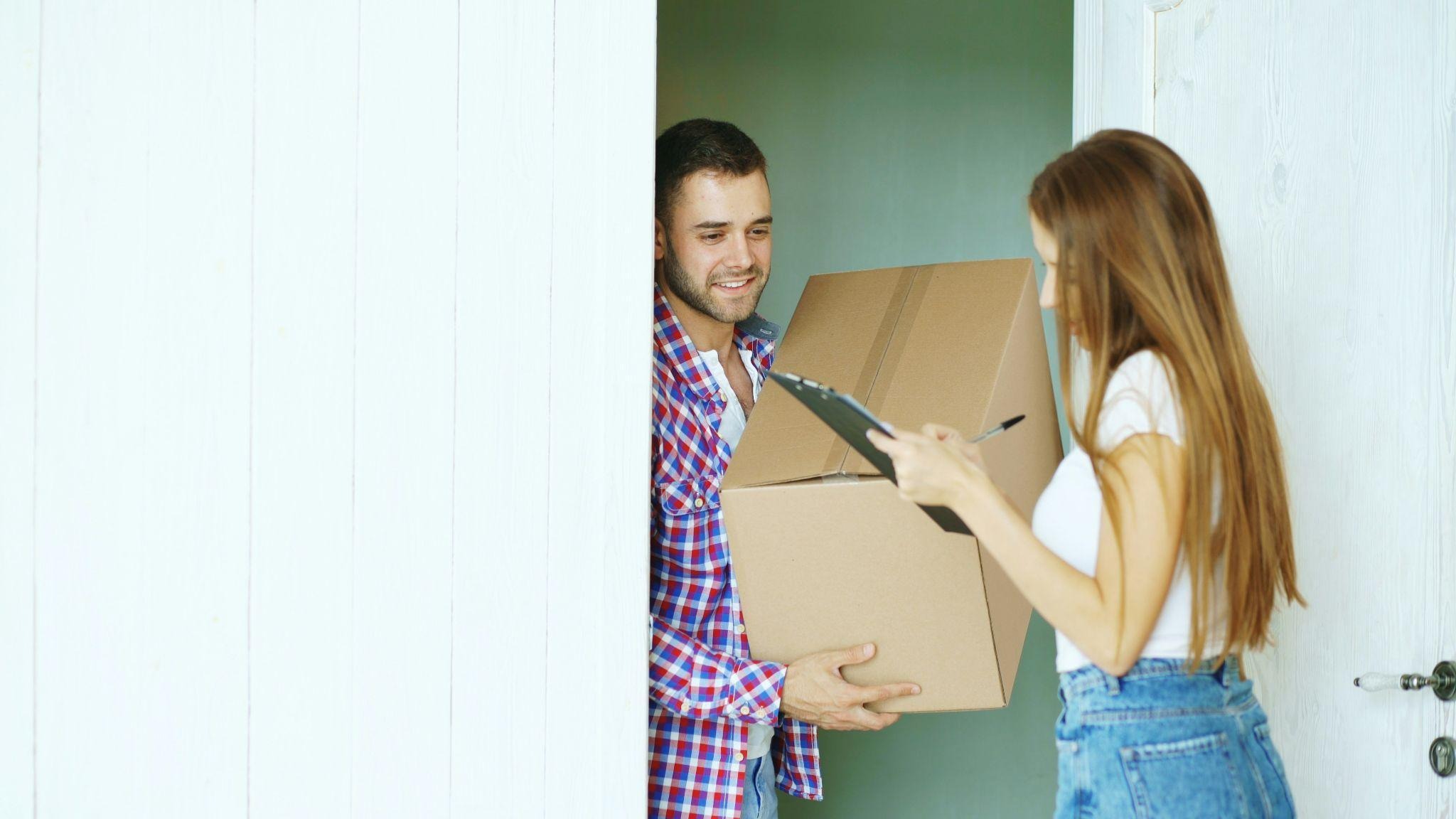 Young couple packing boxes at a home doorway, woman holding a clipboard checklist.